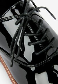 Chaussure en cuir verni noir avec une surface lisse et brillante. Dotée de lacets noirs et d'un détail de couture visible le long des bords.