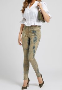 Beige skinny jeans med blå tie-dye mønstre, parret med en hvid bluse med flæser. Outfit'et suppleres af olivengrønne spidse hæle.