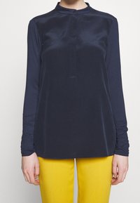 Granatowa bluzka z długim rękawem z gładkiego materiału, z kołnierzykiem mandarynki i jednym guzikiem przy szyi, zestawiona z żółtymi spodniami.