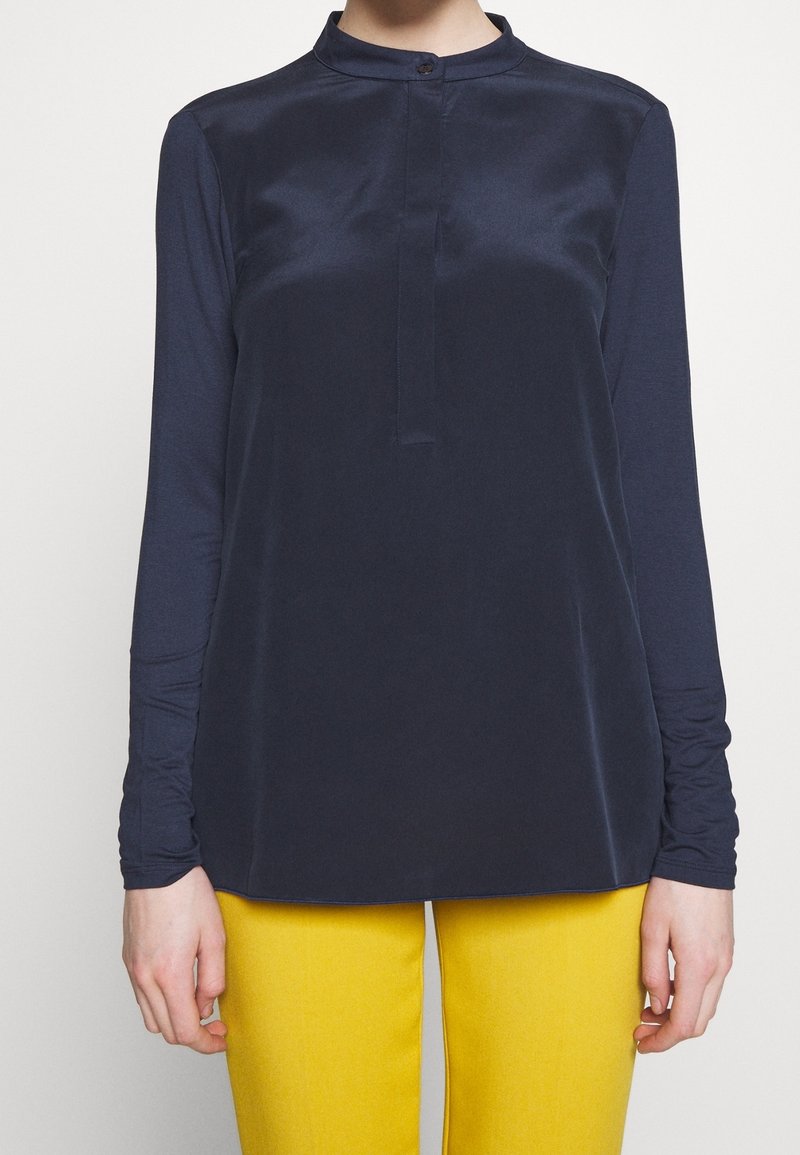 Granatowa bluzka z długim rękawem z gładkiego materiału, z kołnierzykiem mandarynki i jednym guzikiem przy szyi, zestawiona z żółtymi spodniami.