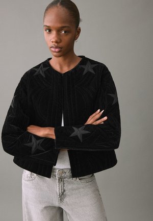 Chaqueta corta de terciopelo negro con diseños de estrellas texturizados y un cuello estructurado. La tela tiene una sensación suave y mullida, con un cierre frontal abierto.