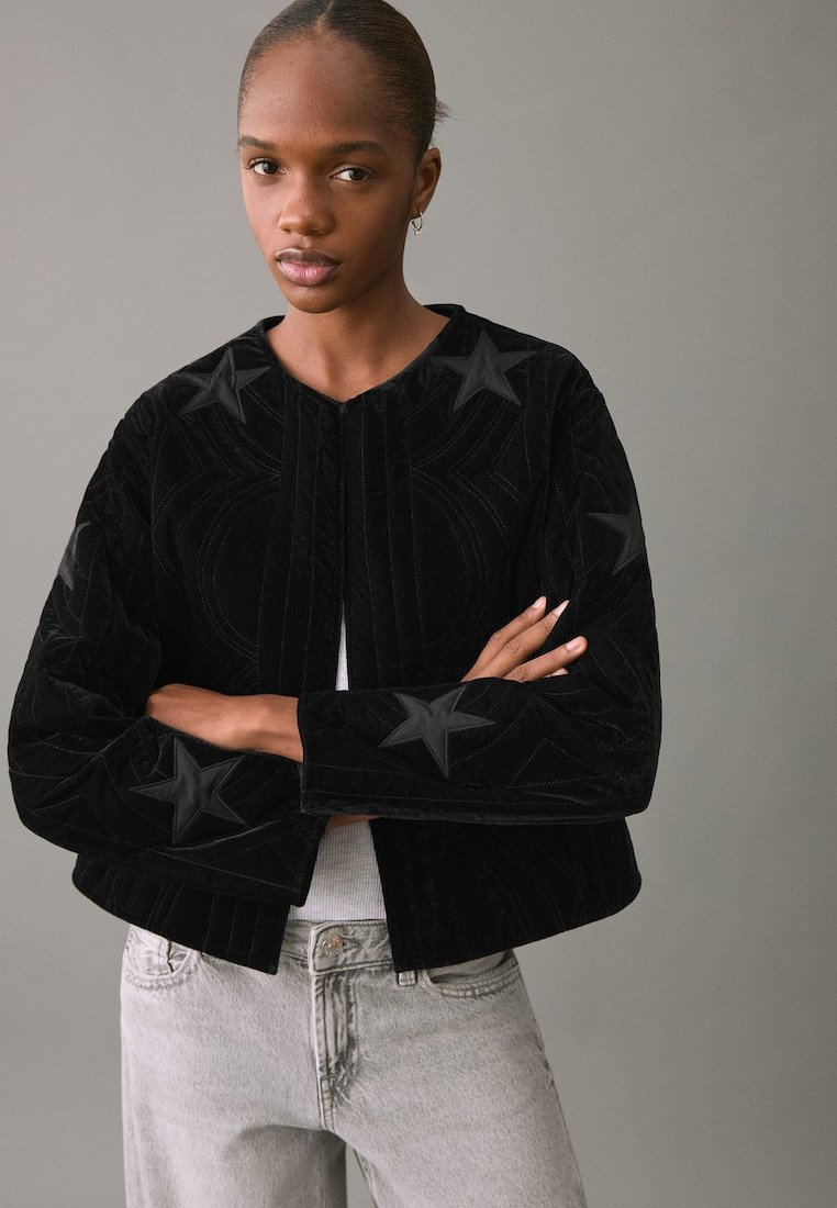 Chaqueta corta de terciopelo negro con diseños de estrellas texturizados y un cuello estructurado. La tela tiene una sensación suave y mullida, con un cierre frontal abierto.