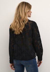 Blusa nera trasparente con motivo floreale in rilievo, maniche lunghe, spalle arricciate e polsini con bottoni. Indossata con jeans blu.