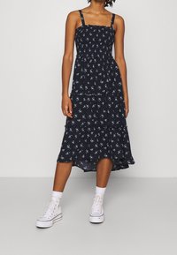 Marinfärgad blommig klänning med figursydd bodice, lager-på-lager kjol och justerbara axelremmar. Bärs med vita högstövlar och strumpor.