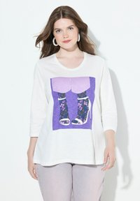 Valkoinen pitkähihainen paita, jossa on violetti graafinen kuva jaloista, jotka ovat kukkakuosisissa sukissa ja korkokengissä. Tausta on yksinkertainen valkoinen.
