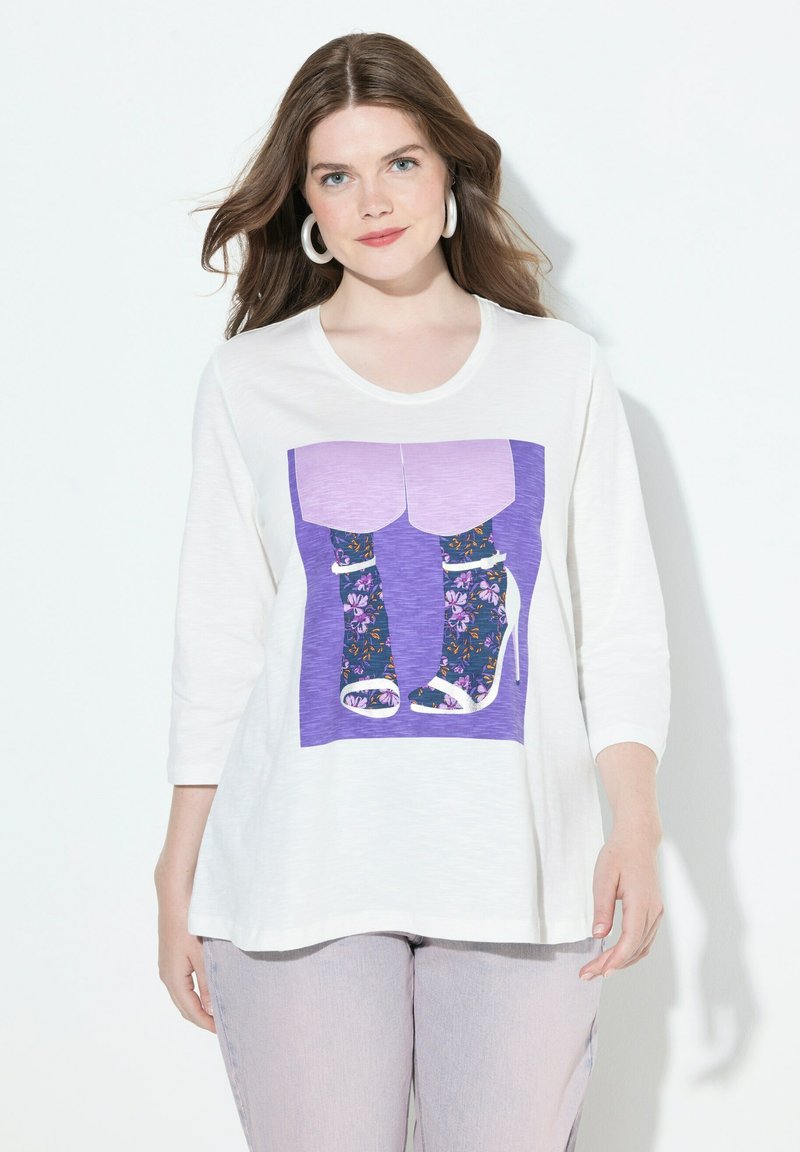 Valkoinen pitkähihainen paita, jossa on violetti graafinen kuva jaloista, jotka ovat kukkakuosisissa sukissa ja korkokengissä. Tausta on yksinkertainen valkoinen.