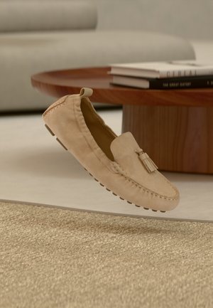 Mocasini din piele întoarsă bej, cu un design cusut, detaliu cu ciucuri și talpă de cauciuc, suspendați deasupra unui covor texturat.