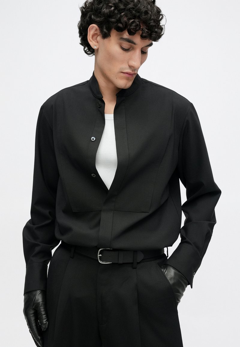 Schwarzes langärmliges Hemd mit hohem Kragen, ausgestattet mit einer vorderen Knopfleiste und einem strukturierten Design. Dazu schwarze Handschuhe und Hose.