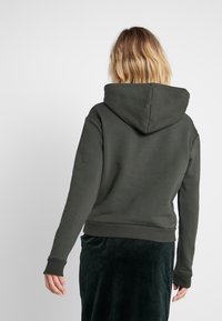 Tmavě zelená mikina s velkou kapucí, žebrovanými manžety a lemem, vyrobená z měkké textilie, navržena pro uvolněný střih. Žádné viditelné vzory ani akcenty.