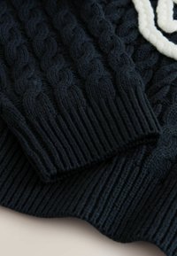 Adīta tumši zila džemperis ar reljefu pinuma rakstu un riboto apmali pie aprocēm. Mīksta tekstūra ar gludu apdari apakšmalā.