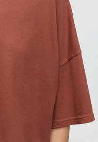 Tissu de chemise à manches courtes de couleur rouille avec couture visible sur la couture de la manche et l'ourlet, porté par une personne sur un fond uni.