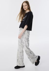 T-shirt noir court avec impression graphique, associé à un pantalon gris comportant des motifs de style manuscrit. Des chaussures noires complètent la tenue.