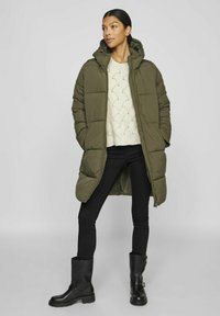 Vadderad lång kappa i olivgrönt med huva, bärs över en tröja i krämfärgad stickning och svarta tajta jeans, kombinerat med svarta ankelstövlar.