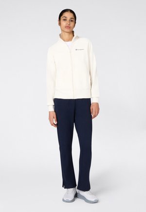 Donna in piedi, veste una giacca bianca con zip, pantaloni blu navy e sneakers bianche su uno sfondo chiaro uniforme.