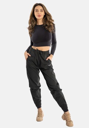 Schwarzes, kurz geschnittenes Langarmoberteil und lockere schwarze Hose mit elastischen Bündchen, kombiniert mit hellbraunen Schnürschuhen. Das Oberteil hat einen Kordelzug am Bund.