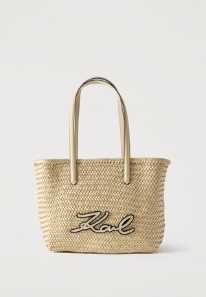Υφαντή τσάντα από ψάθα με μπεζ δερμάτινες χειρολαβές και λογότυπο «Karl» σε καλλιγραφικό στυλ με μαύρο περίγραμμα στο μπροστινό μέρος, σε απλό λευκό φόντο.