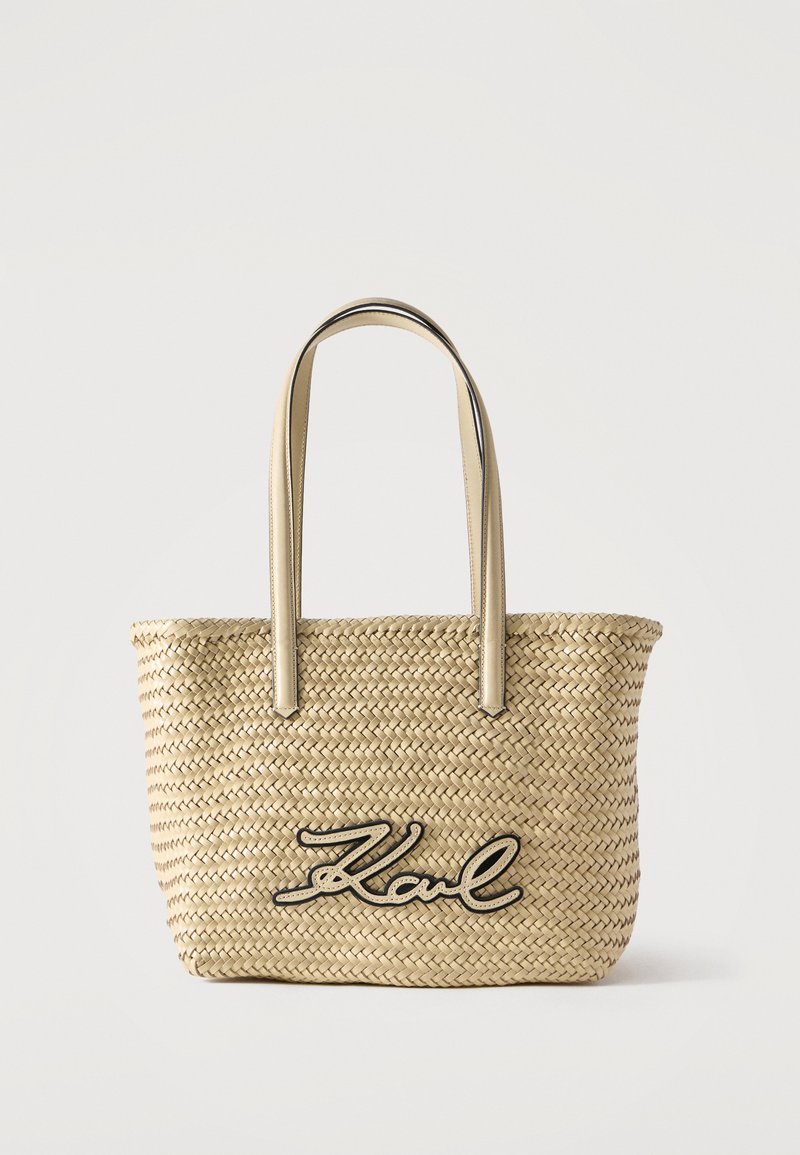 Υφαντή τσάντα από ψάθα με μπεζ δερμάτινες χειρολαβές και λογότυπο «Karl» σε καλλιγραφικό στυλ με μαύρο περίγραμμα στο μπροστινό μέρος, σε απλό λευκό φόντο.
