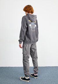 Harmaa huppari, jossa on palmukuvio ja teksti selässä, yhdistettynä vaaleanharmaisiin cargo-housuihin ja mustiin tennareihin. Puuvillamateriaali, rento muotoilu.