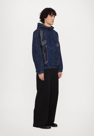 Sweat à capuche bleu marine avec un motif texturé et des motifs en fil clair contrastant. Coupe décontractée, associé à un pantalon noir large et des chaussures lisses.