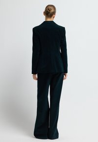 Žena s vlasy v drdolu, oblečená v tmavě tyrkysovém sametovém obleku, stojící zády k fotografovi na bílém pozadí.