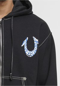Černá mikina s kapucí na zip, která má bílé švy a modrý grafický logotyp s nápisem "TRUE RELIGION." Látka vypadá jemně a má hladkou strukturu.