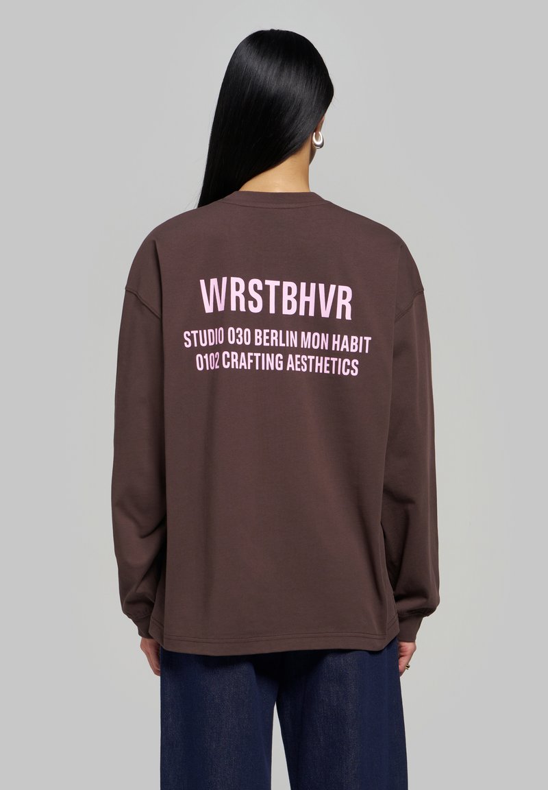 Femme aux longs cheveux noirs portant un sweat-shirt marron avec un texte rose "WRSTBHVR" et d'autres mots, debout devant un fond gris uni.