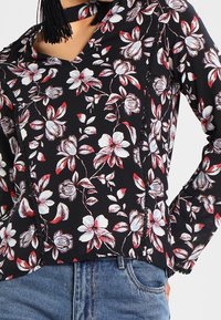 Blouse noire à fleurs rouges et blanches, avec un col en V et des bords volantés. Associée à un jean en denim bleu.