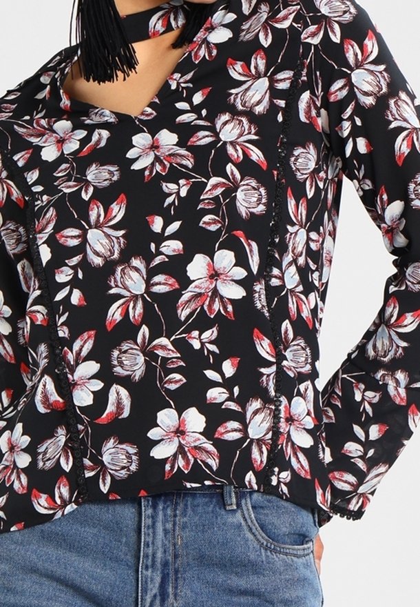 Blouse noire à fleurs rouges et blanches, avec un col en V et des bords volantés. Associée à un jean en denim bleu.