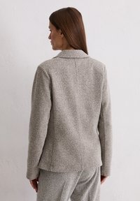 Blazer in light gray textured fabric with a notch collar, single back vent, and long sleeves, shown from the back.