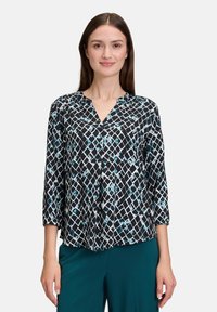 Blouse à motifs noir et blanc avec des manches trois-quarts et un col en V. Présente un design géométrique avec des accents de turquoise et de blanc.