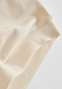 Beige Sweatshirt-Detail mit einer glatten, weichen Textur und einem gerippten Saum. Der Stoff ist leicht und scheint sich gut für Freizeitkleidung zu eignen.