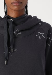 Sudadera negra con cuello en cowl, que presenta patrones de estrellas blancas y cordones. Textura suave con acentos metálicos en las puntas de los cordones.