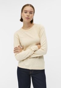 Pull beige à manches longues avec col rond et poignets côtelés, fabriqué en matériau doux, associé à un jean bleu foncé. Design simple et polyvalent.