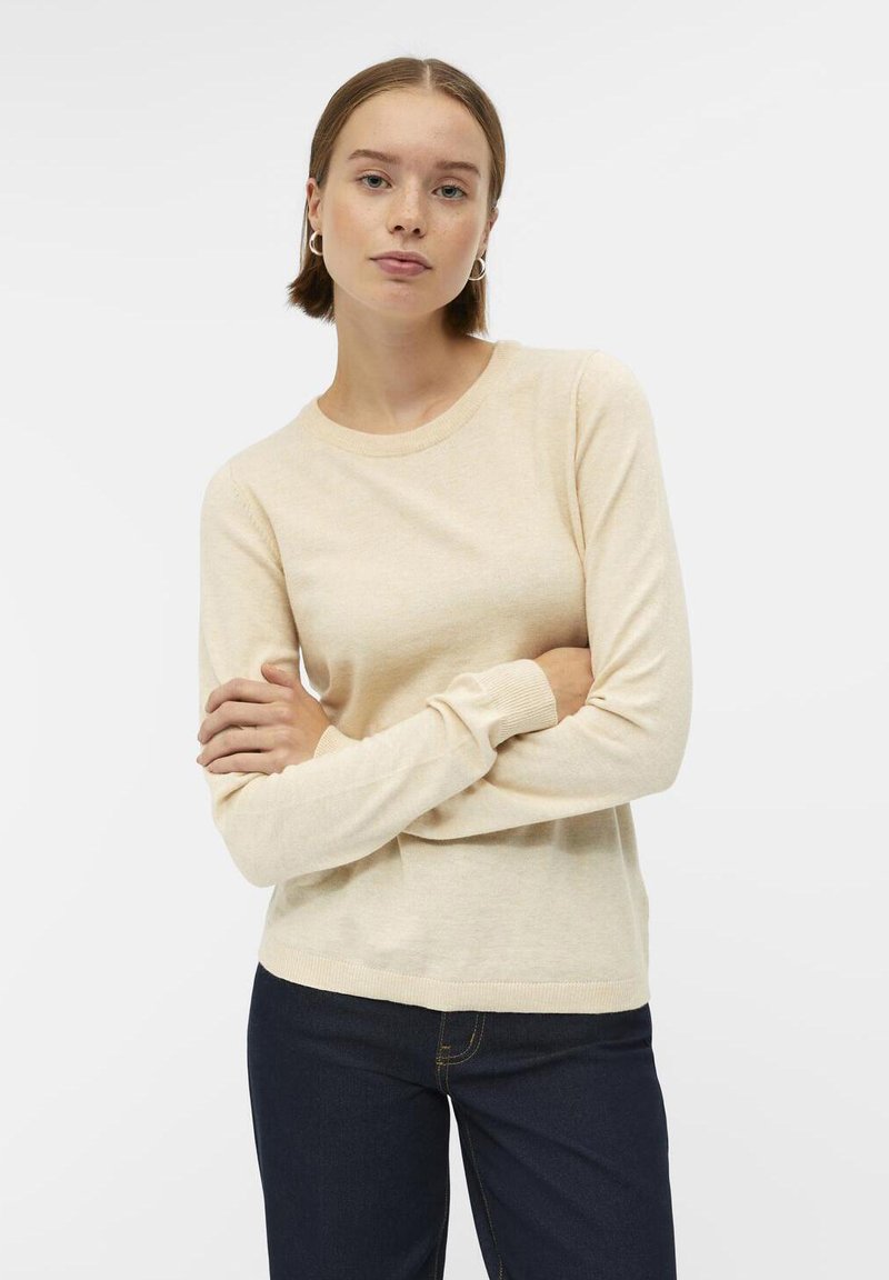 Pull beige à manches longues avec col rond et poignets côtelés, fabriqué en matériau doux, associé à un jean bleu foncé. Design simple et polyvalent.