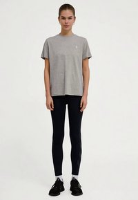 Graues Baumwoll-T-Shirt mit Rundhalsausschnitt und kurzen Ärmeln, kombiniert mit schwarzen Leggings mit seitlichem Muster, getragen mit schwarzen Turnschuhen.