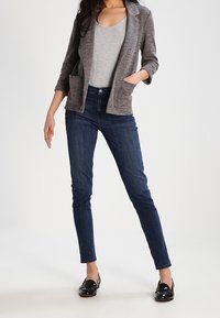 Szary fakturowany blezer z dwoma przednimi kieszeniami, w połączeniu z dopasowanymi ciemnoniebieskimi dżinsami i czarnymi loafersami, noszony na szarym topie.