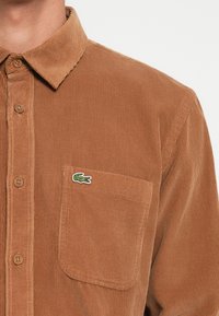Brown corduroy shirt with a button-down collar, front pocket, and a green embroidered crocodile logo. Textured, ribbed fabric detail visible.
