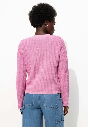 Pull en tricot rose avec une texture côtelée et des manches longues, présentant un col rond et un ourlet ajusté, associé à un pantalon cargo bleu.