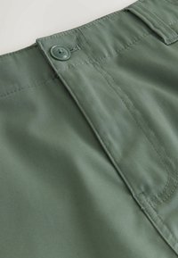 Gros plan sur la ceinture d'un pantalon vert olive avec fermeture par bouton, passants pour ceinture et coutures détaillées le long de la braguette et de la ceinture.