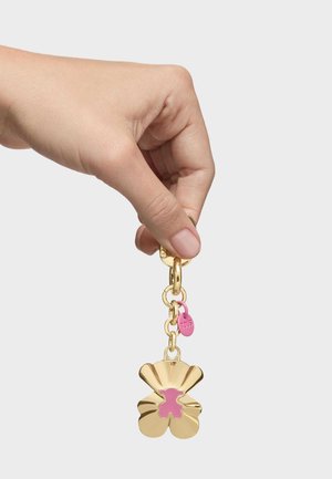 Mano che tiene un portachiavi dorato con un ciondolo a forma di orsetto smaltato di rosa e una piccola targhetta rosa con la scritta "TOUS" su sfondo bianco.
