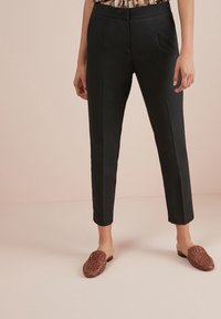 Czarne spodnie o prostym kroju, wykonane z gładkiego materiału i subtelnych fałd, w połączeniu z brązowymi plecionymi butami wsuwanymi.