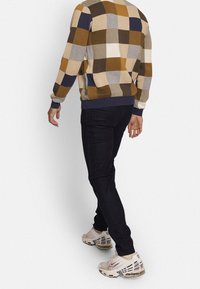 Pull en motif carreaux marron, bleu marine et crème avec tricot texturé ; associé à un jean foncé et des baskets blanches.