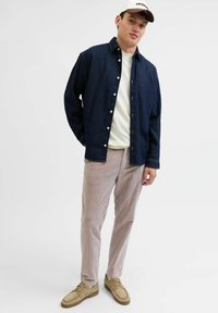 En mørk denim skjorte med knapper, en hvid t-shirt, lyse stribede fløjlsbukser og beige sko, båret af en person der står afslappet.