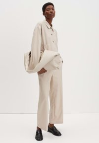 Camicia giacca oversize beige con grandi bottoni, pantaloni larghi abbinati e mocassini neri; tessuto texture, design minimalista.