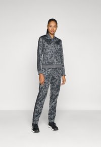 Grå leopardmönstrad träningsdräkt med zip-up jacka och matchande byxor, tillverkad av mjukt material. Har svarta ränder längs sidorna.