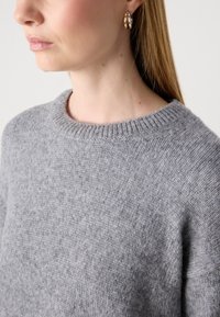 Mulher a usar um suave suéter de malha cinza e brincos em forma de argola dourados, mostrada do pescoço para baixo com cabelo longo e liso em fundo simples.