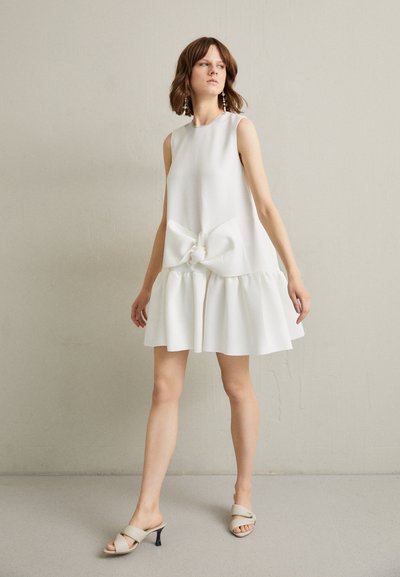 Bijela haljina bez rukava s velikom mašnom na struku i lepršavim volanima na rubu. Model je kombinirao s bež sandalama na petu.