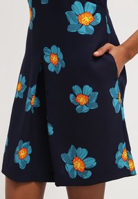 Vestido de marino con grandes flores turquesa y centros naranjas. La tela es suave y tiene un ligero brillo; incluye bolsillos laterales.