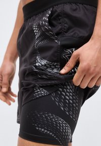 Κοντινό πλάνο ενός ατόμου που φοράει μαύρα αθλητικά σορτς με γκρι μοτίβο φολίδων φιδιού, προσαρμόζοντας το ύφασμα της πλαϊνής τσέπης.