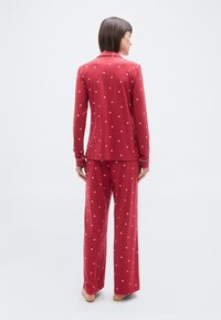 Femme debout pieds nus de dos, portant un pyjama rouge à manches longues avec de petits motifs de cœurs blancs et un liseré blanc sur le col et les poignets.